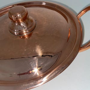 May include: A copper pot with a lid and two handles. The pot has a hammered finish and a shiny surface.