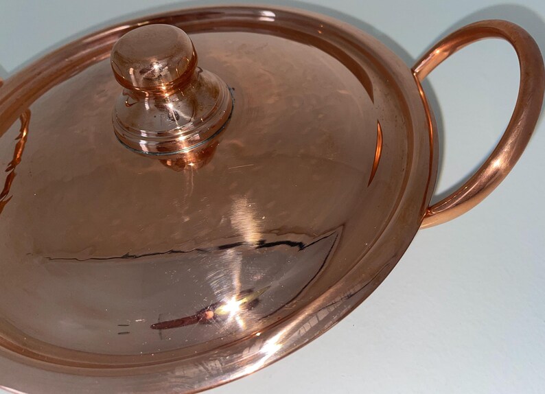 May include: A copper pot with a lid and two handles. The pot has a hammered finish and a shiny surface.