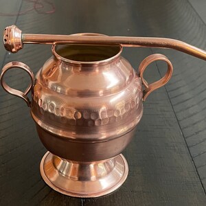 Puede incluir: Una tetera de cobre con dos asas y un acabado martillado. Tiene un pico en la parte superior y una tapa de cobre a juego.