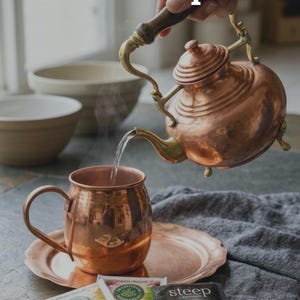 May include: A copper tea set with a teapot pouring water into a mug. The set includes a saucer, and tea packets. The text reads "Confidence in the Prep." and "Naturally antibacterial copper protects your family while elevating your daily tea."