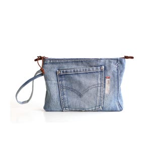 Op de afbeelding: Een lichtblauw denim tasje met een voorvak met V-vormige stiksels, een metalen ritssluiting met een bruin leren treklipje en een bijpassende denim polsband. De stof heeft een vervaagde wassing met gerafelde randen en een klein rood label.