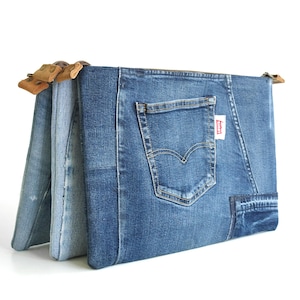 May include: A denim clutch made from recycled jeans. The clutch features a back pocket and a small red tag. The clutch is a patchwork of different shades of blue denim and has a wooden handle.