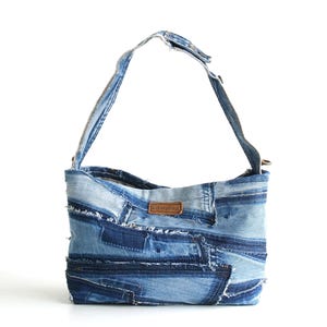 Op de afbeelding: Een blauwe denim schoudertas gemaakt van gerecyclede jeans. De tas heeft een patchwork ontwerp met rafelige randen en een bruin lederen label.