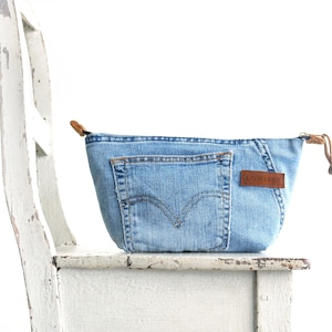 May include: A light blue denim pouch with a brown leather tag and zipper pull. The pouch features a back pocket and is sitting on a white distressed chair. The tag reads "LOWE".