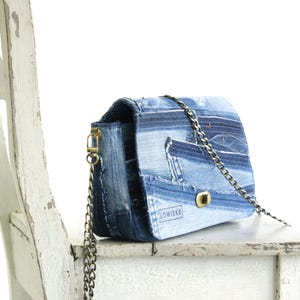 Op de afbeelding: Een klein blauw denim schoudertasje met een gouden kettingband. De tas is gemaakt van gerecycled denim en heeft een patchwork ontwerp. De tas heeft een klein gouden sluiting en het woord "LOWIEKE" is op de voorkant gedrukt.