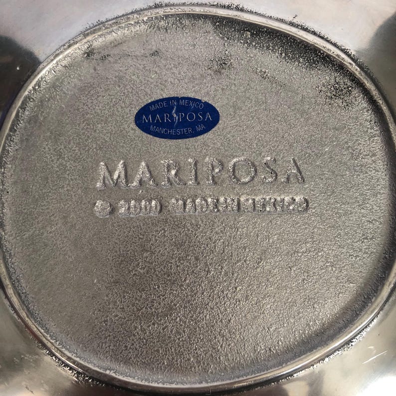 May include: Close-up of a silver-toned metal object with the embossed word "MARIPOSA." A blue oval sticker reads "MADE IN MEXICO MARIPOSA MANCHESTER, MA." The object has a textured surface and a circular shape.