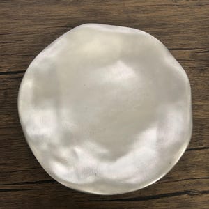 May include: A silver-colored, irregularly shaped plate with a smooth, reflective surface. The plate has a slightly uneven rim, giving it a handcrafted appearance. The plate is likely made of metal and is suitable for serving food.