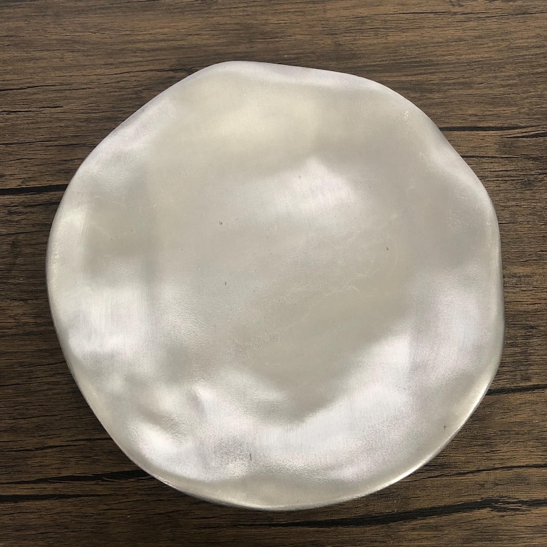 May include: A silver-colored, irregularly shaped plate with a smooth, reflective surface. The plate has a slightly uneven rim, giving it a handcrafted appearance. The plate is likely made of metal and is suitable for serving food.