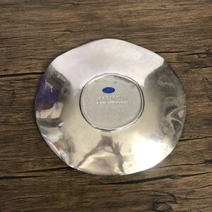 May include: A silver-colored, irregularly shaped decorative plate. The plate has a smooth, reflective surface with a slightly hammered texture. The center of the plate features a circular area with the word "MARIPOSA" and "MADE IN MEXICO" printed on it.