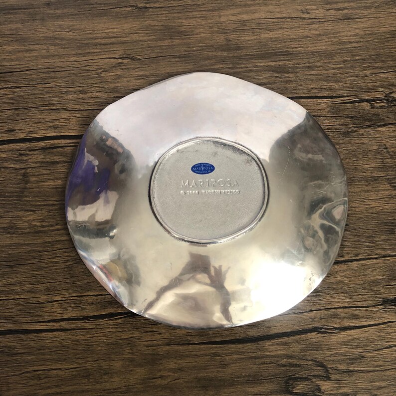 May include: A silver-colored, irregularly shaped decorative plate. The plate has a smooth, reflective surface with a slightly hammered texture. The center of the plate features a circular area with the word "MARIPOSA" and "MADE IN MEXICO" printed on it.