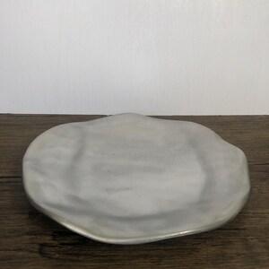 May include: A light gray, organically shaped ceramic serving dish. The dish has a smooth, matte finish and a slightly raised edge. The dish is sitting on a wooden surface.