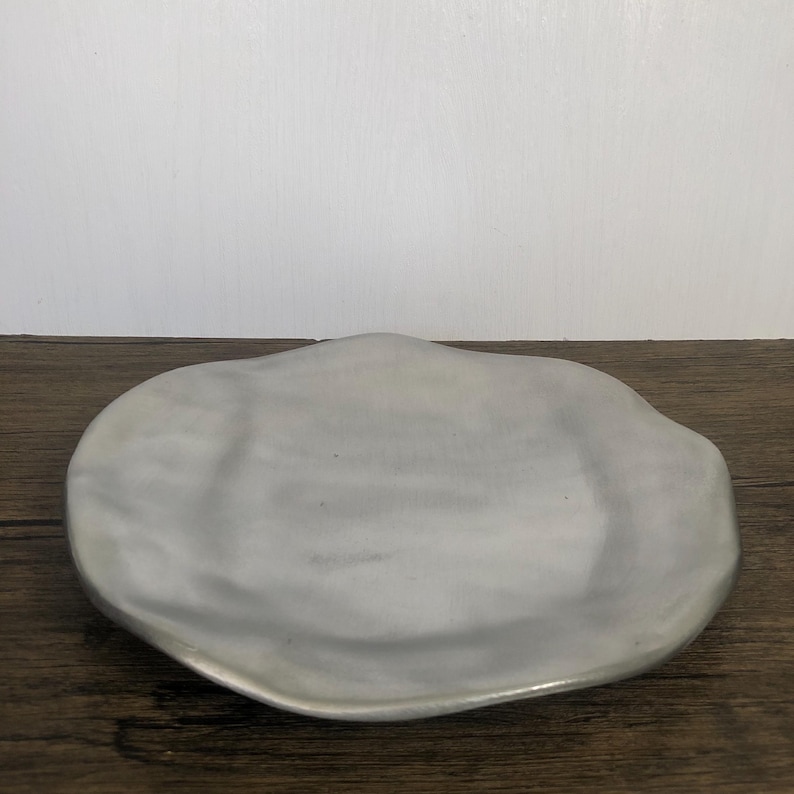 May include: A light gray, organically shaped ceramic serving dish. The dish has a smooth, matte finish and a slightly raised edge. The dish is sitting on a wooden surface.