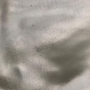 May include: Close-up of a textured, silver-colored surface. The surface has a slightly rough, grainy appearance with subtle variations in tone and small dark specks.