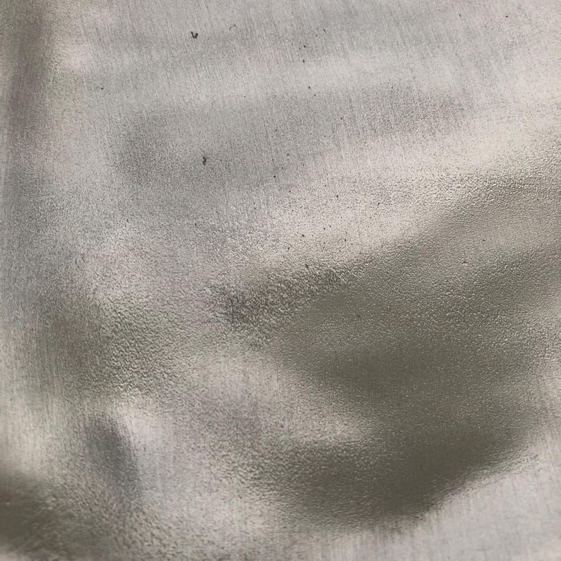 May include: Close-up of a textured, silver-colored surface. The surface has a slightly rough, grainy appearance with subtle variations in tone and small dark specks.