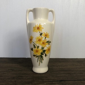 Puede incluir: Jarrón de cerámica color crema con dos asas y diseño floral. El jarrón presenta un ramo de margaritas amarillas con hojas verdes. El jarrón mide aproximadamente 25 cm de alto, con base redondeada y parte superior acampanada.