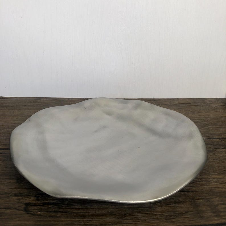 May include: A silver-colored, irregularly shaped decorative plate. The plate has a smooth surface and a slightly raised edge. The plate is sitting on a dark wooden surface.