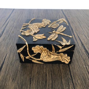 May include: Black square jewelry box with gold-colored floral and dragonfly relief carvings. The box features a textured surface and is set on a wooden surface.