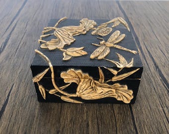 May include: Black square jewelry box with gold-colored floral and dragonfly relief carvings. The box features a textured surface and is set on a wooden surface.