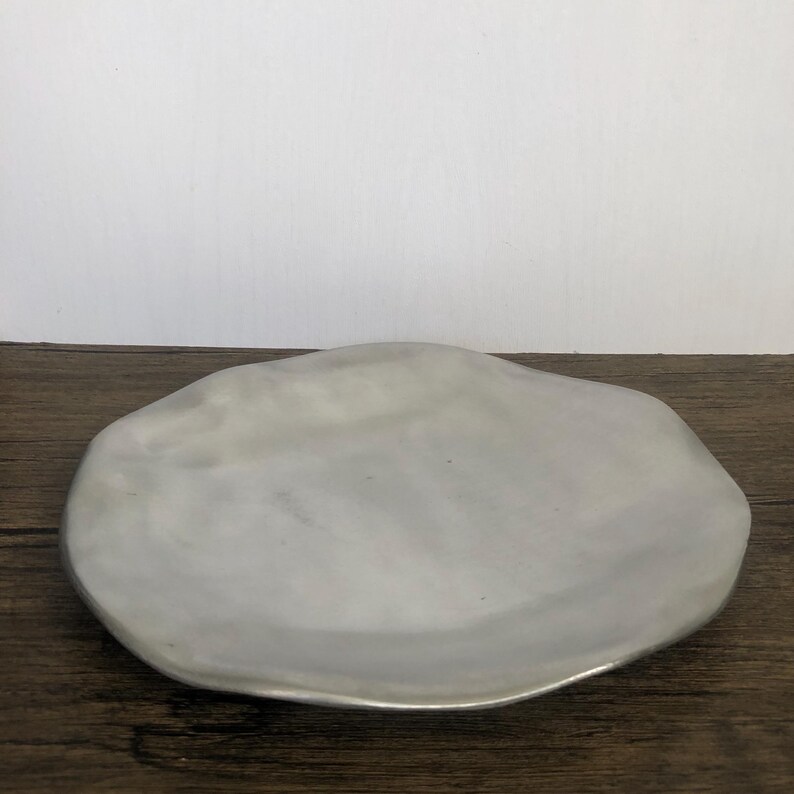 May include: A silver-colored, organically shaped plate. The plate has a smooth surface and a slightly raised edge. The plate is sitting on a dark wooden surface. The plate is a decorative item.