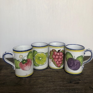 May include: Four ceramic mugs with fruit designs. Each mug features a different fruit: a strawberry, a lime, grapes, and a plum. The mugs have blue and yellow trim and patterned handles.