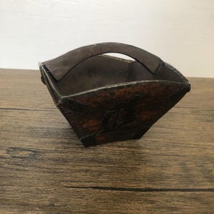 May include: A dark brown, handcrafted basket with a handle. The basket has a square shape with a decorative design on the front. The handle is curved and attached to the top of the basket. The basket is made of a dark, possibly aged, material.