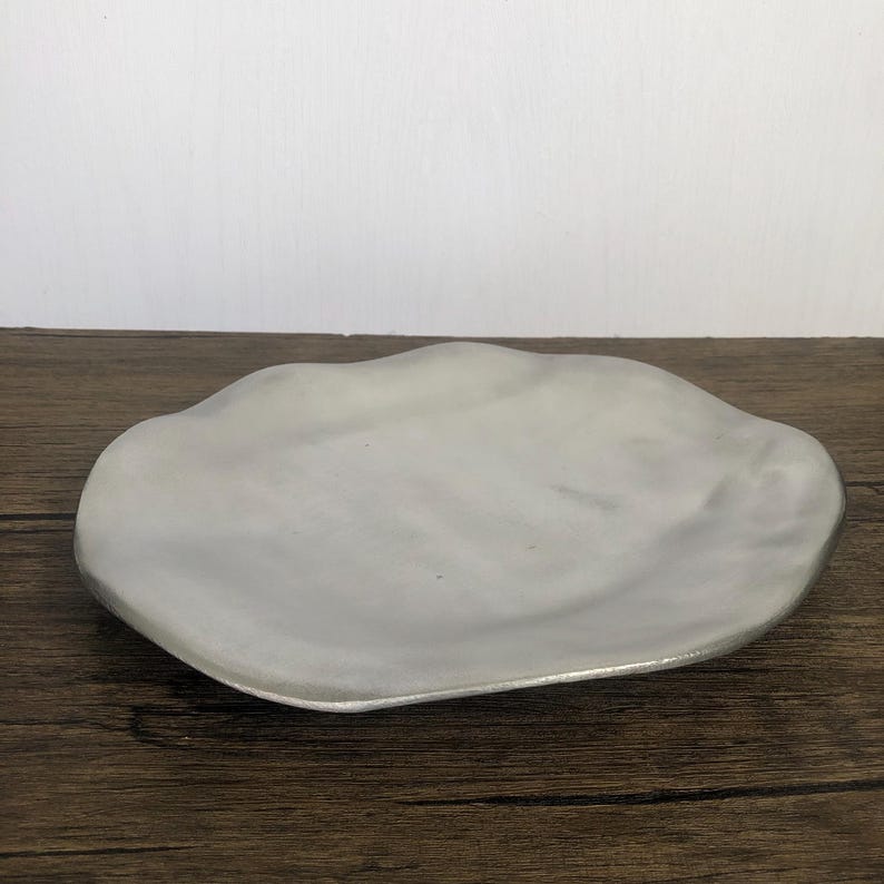 May include: A decorative, irregularly shaped, silver-colored plate. The plate has a wavy edge and a smooth, reflective surface. The plate is made of metal and is suitable for serving food or as a decorative item. The plate is on a wooden surface.