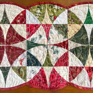 May include: A quilted Christmas table runner with a scalloped edge. The runner features a repeating pattern of red, green, and white fabric sections. The design includes star-like shapes and floral patterns, creating a festive holiday theme.