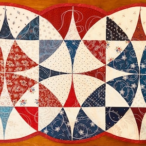May include: A patriotic-themed quilted table runner with a scalloped edge. The runner features a patchwork design with red, white, and blue fabrics. The pattern includes floral and star motifs, creating a festive and decorative piece, approximately 40 inches long.