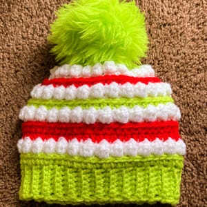 May include: A green and red striped crocheted hat with a white pom-pom. The hat has a green crocheted brim.