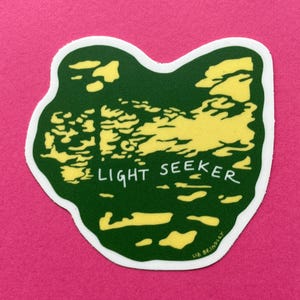 May include: A die-cut sticker with a green and yellow landscape design. The sticker features the text "LIGHT SEEKER" in white. The sticker is on a pink background.