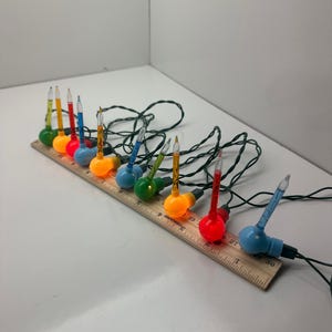 May include: A string of multi-colored bubble Christmas lights with green wire, featuring round bases in red, blue, green, yellow, and orange, positioned along a wooden ruler measuring approximately 12 inches in total length.