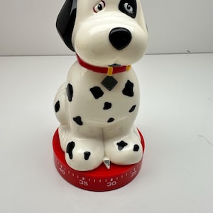 May include: A white kitchen timer shaped like a sitting dog with black spots, a black ear, and a black nose. The dog wears a red collar with a small gray tag. The base is a circular red dial featuring white numerical markings for time intervals.