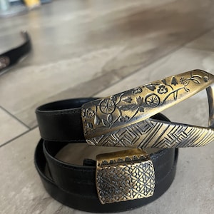 May include: Black leather belt with ornate gold-colored buckles. The larger buckle features a floral and geometric design, while the smaller buckle has a repeating pattern. The belt is coiled, showcasing its length and design.