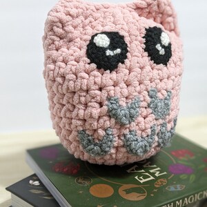 May include: A pink crocheted owl with black eyes and a grey beak, sitting on top of two books. The top book has a green cover with the text "Earth Magic" and the bottom book has a black cover.