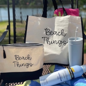 May include: Two white canvas tote bags with black trim and the text "Beach Things" in black script. A smaller white canvas pouch with black trim and the text "Beach Things" in black script. A blue tumbler with a straw and a bottle of sunscreen.