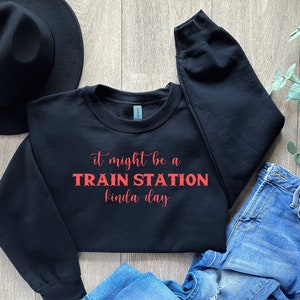 May include: A black long-sleeve crewneck sweatshirt featuring the red text "it might be a TRAIN STATION kinda day" printed on the front. The garment has a ribbed collar, cuffs, and hem, and is displayed alongside a black hat and blue jeans.