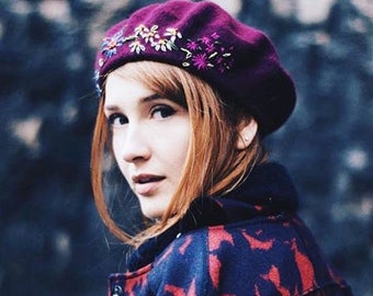 May include: A woman wearing a purple beret with embroidered flowers. The beret is made of felt and has a soft, comfortable texture.