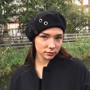 Puede incluir: Un gorro negro de estilo beret con ojales plateados. El gorro lo lleva puesto una persona con el pelo largo y castaño.