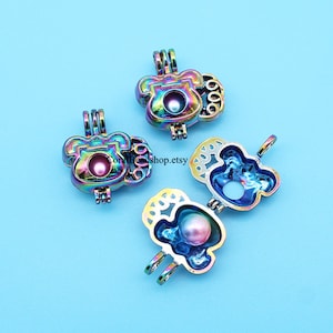 May include: Three iridescent metal charm pendants with a pearl inside. The charms are shaped like a telephone, a bear, and a bear with a crown. The charms are open and the pearls are visible.