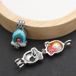 May include: Two silver charm pendants with a rainbow and a sunset design. The charms are open and have a small pearl inside. The charms are on a wooden surface.