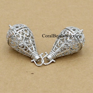 Puede incluir: Dos colgantes de jaula de filigrana plateados con un diseño de corazón. Los colgantes tienen un bucle en la parte superior para sujetarlos a una cadena o cordón.