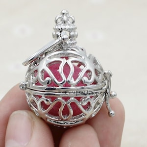 Puede incluir: Colgante de jaula de filigrana plateada con interior rosa. La jaula está abierta y tiene una pequeña parte superior en forma de corona.