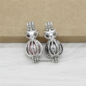 May include: Two silver-toned metal owl-shaped bead cages, each featuring a vertical wire design. The left cage holds a round, light pink bead, while the right cage contains a round, grey bead. The text CoralBeadshop.etsy is visible at the bottom.
