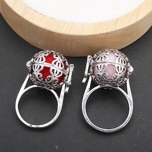 Op de afbeelding: Twee zilveren ringen met een ronde, opengewerkte kooi die een kleine bal bevat. Eén bal is rood en de andere is roze.