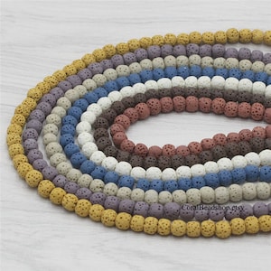 Può includere: Una collezione di dieci fili di perline di roccia vulcanica in vari colori, tra cui giallo, beige, grigio, viola, blu e marrone. Le perline sono rotonde e hanno una texture ruvida e porosa.