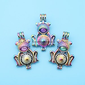 Puede incluir: Tres colgantes iridiscentes con forma de rana y un diseño floral en el vientre. Los colgantes tienen un acabado metálico arcoíris y están sobre un fondo azul claro. Los colgantes son probablemente para la fabricación de joyas.