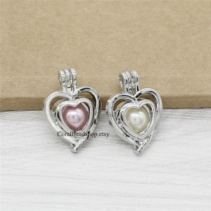 May include: Two silver heart-shaped locket pendants with a pearl inside each. The pearl in the left pendant is pink, and the pearl in the right pendant is white.