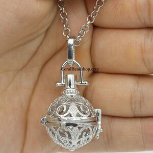 May include: A silver-toned metal locket pendant featuring an intricate filigree pattern with a spherical shape. The pendant is attached to a silver-toned link chain and includes a small bail at the top. The text CoralBeadshop.etsy is visible.