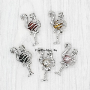 May include: Five silver flamingo charm cages with different colored pearls inside. The cages are open on the top and bottom, and the pearls are visible. The cages are hanging from a white surface.