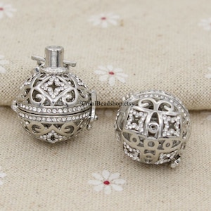 Puede incluir: Dos abalorios de bola de jaula de metal plateado con acentos de cristal transparente. Los abalorios están abiertos y tienen un diseño de filigrana.